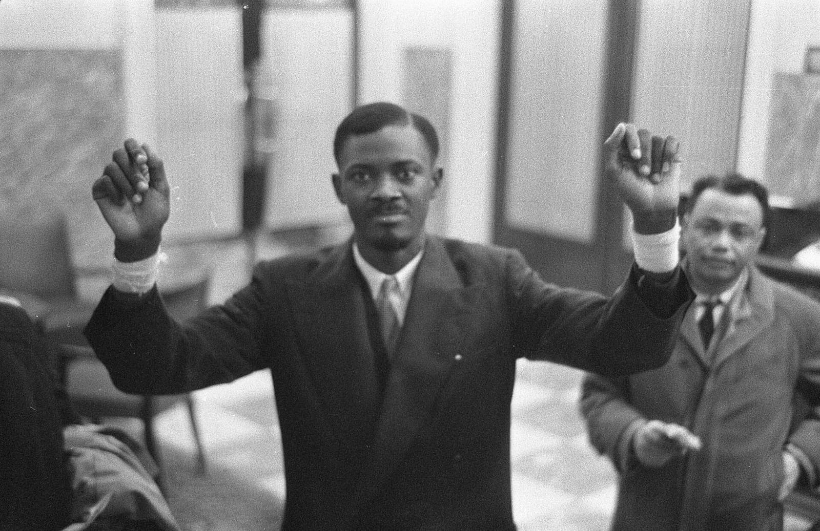 Patrice Lumumba standing with both arms raised, addressing a room.